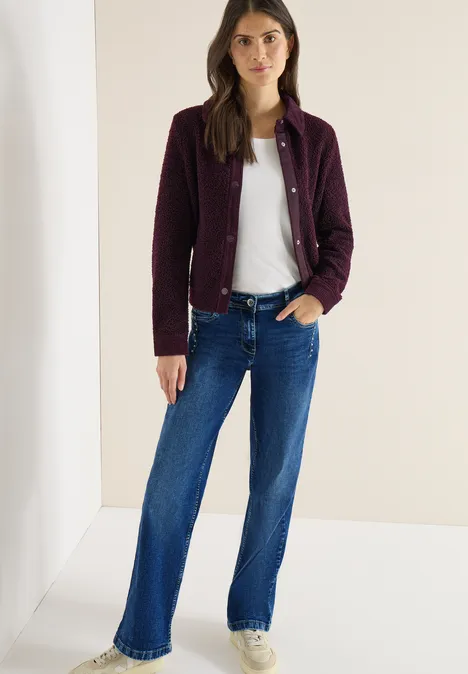 Frau im violetten Cardigan, weißem T-Shirt und blauen, ausgestellten Jeans.